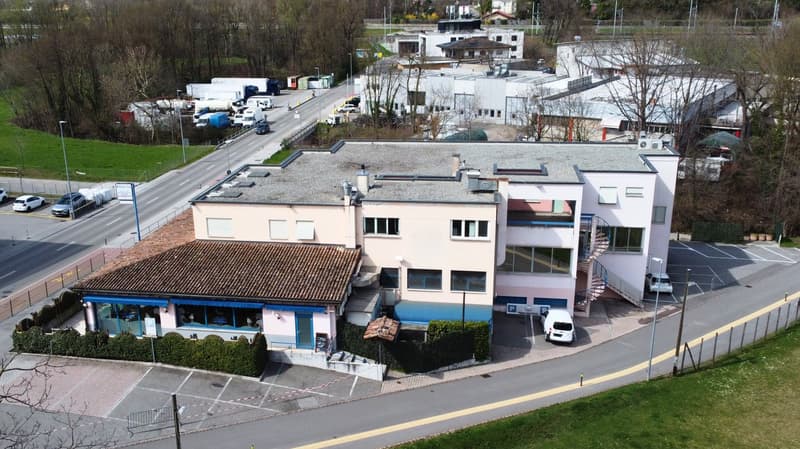 The image shows a commercial/industrial area with several buildings and structures. There is a road running through the area with parked vehicles visible. The buildings appear to be a mix of commercial and industrial structures, with some having a more mo
