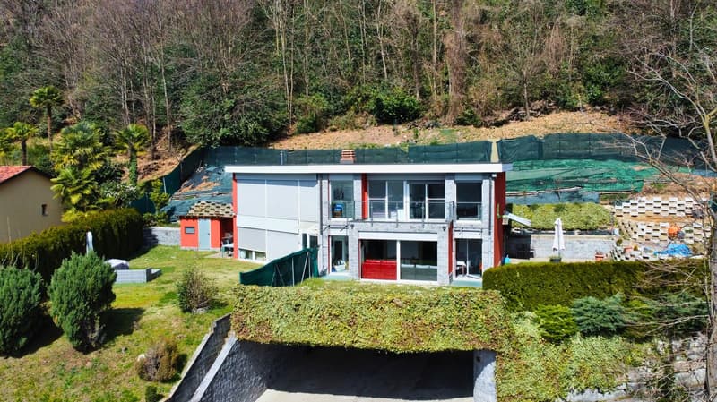 Two-story modern building with large windows, balcony, and surrounded by lush greenery and trees