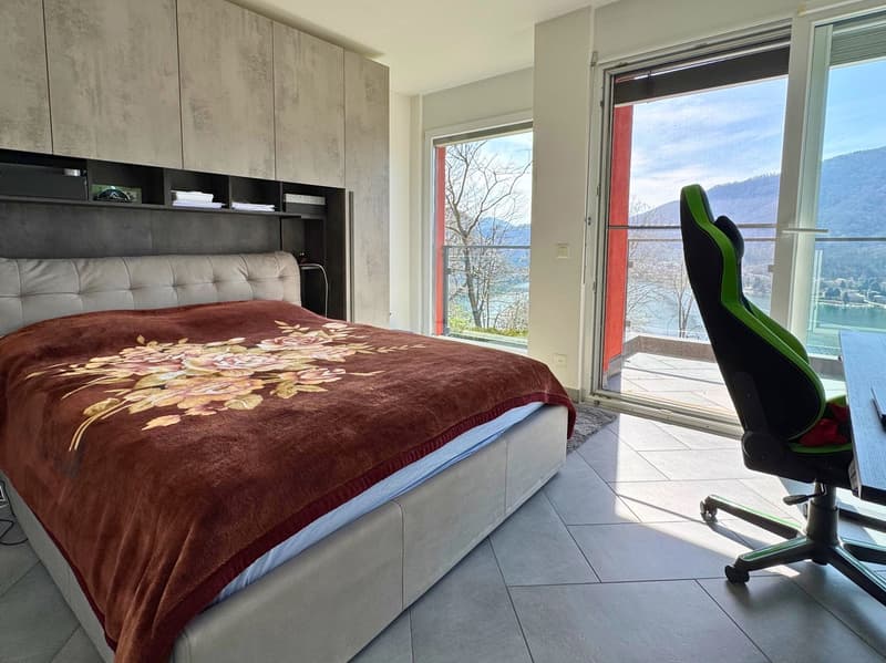 Spacious bedroom with large windows offering a scenic view of the surrounding landscape. The room features a plush, floral-patterned bedspread, built-in storage cabinets, and a comfortable-looking gaming chair positioned near the window.