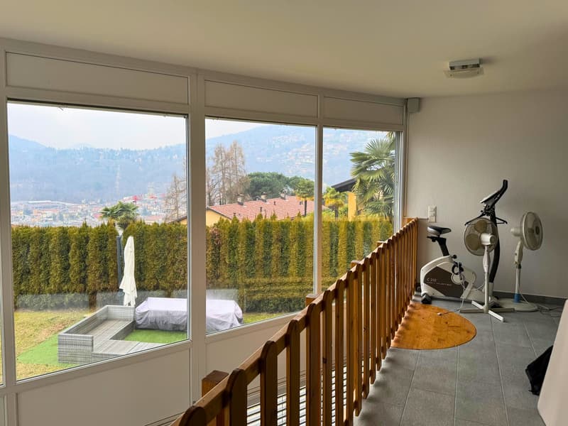 Large windows with a view of the surrounding mountains and buildings, wooden balcony railing, exercise equipment such as a stationary bike, and a covered outdoor seating area visible through the windows.