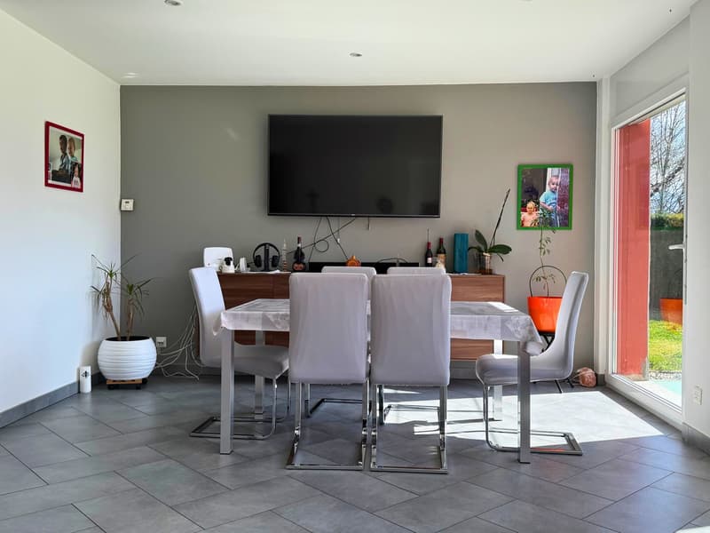 Spacious dining area with a large wooden dining table and white chairs, a mounted TV on the wall, and a sideboard with various decor items. The room has gray walls and tile flooring, and a sliding glass door leading to an outdoor area.