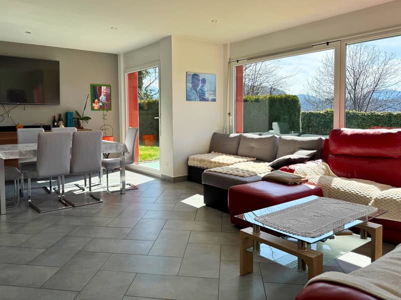 Spacious open-plan living area with large windows overlooking a garden, red leather sofa, gray sectional sofa, glass coffee table, dining table and chairs, tiled flooring, artwork on the walls