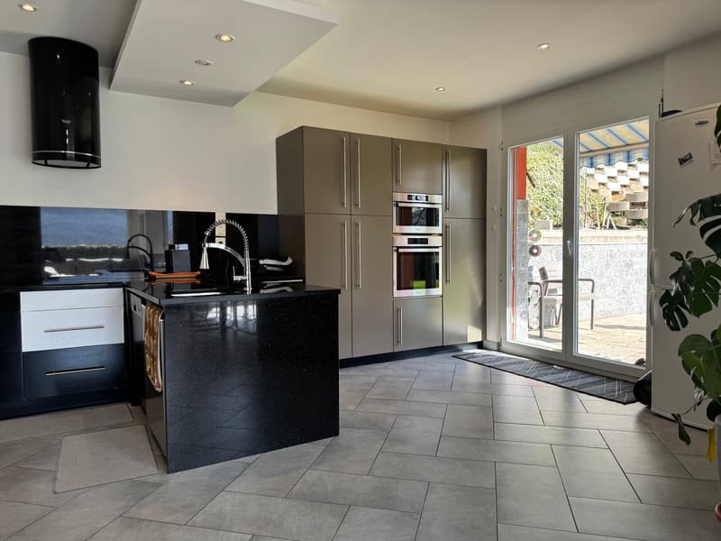 Modern kitchen with grey cabinets, black countertop, built-in appliances including dishwasher and washing machine, large windows providing natural light