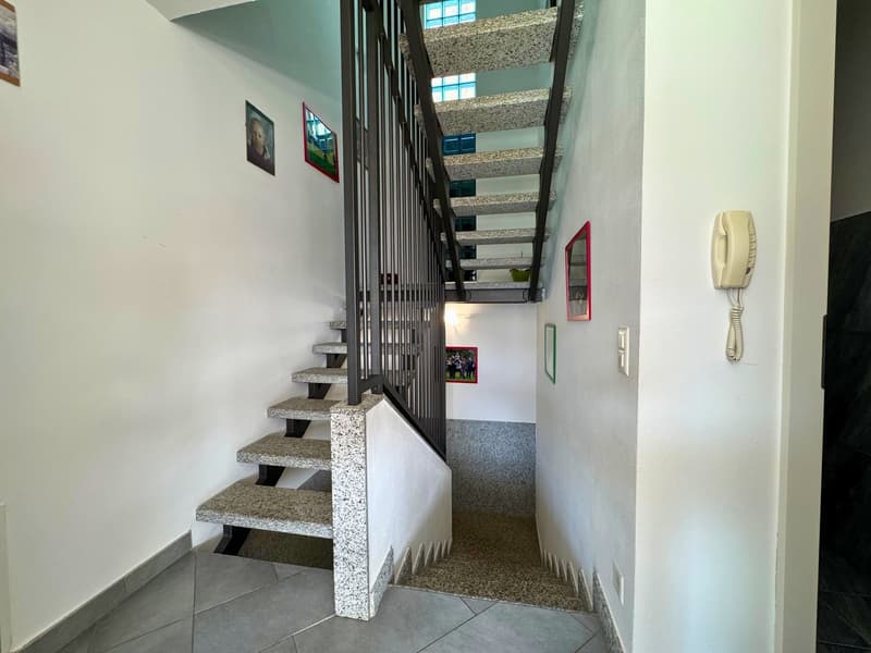 The image shows a staircase with granite steps and a metal railing. The walls are painted in a light color and there are some framed pictures hanging on the walls. The staircase appears to be the main entrance to the building.
