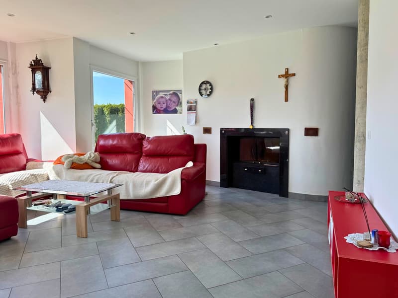 Spacious living room with red leather sofa, coffee table, wall-mounted TV, religious decor such as a cross and framed photo, and tiled flooring.