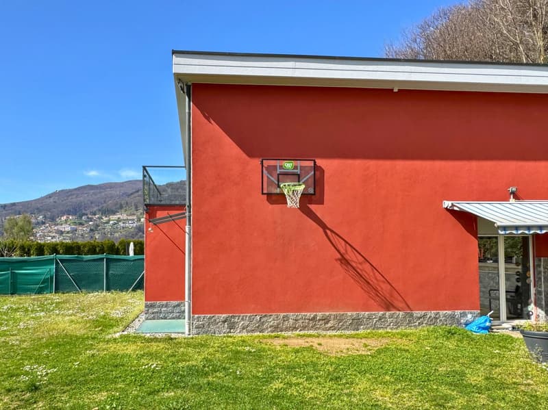 Two-story building with a red exterior, basketball hoop, and a grassy area in front. The building appears to be located in a mountainous area with a blue sky in the background.
