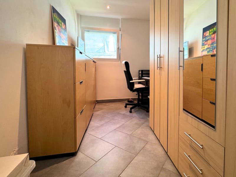 The image shows a small office space with a desk, chair, and various storage cabinets. The room has tile flooring and wooden cabinetry. There is a window visible, and the walls are painted a light color. The room appears to have a washing machine and dish