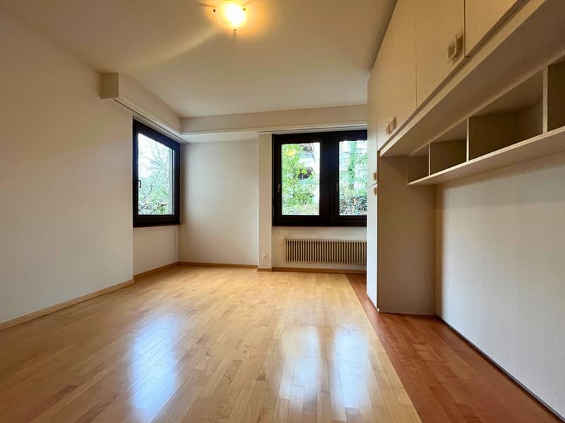 large wooden floor, white walls, 2 windows, overhead light, radiator, ceiling cabinets, light switch