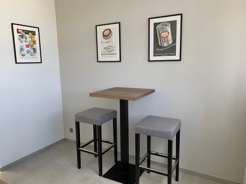 Small kitchen area with a wooden table and two gray bar stools. The walls feature framed vintage advertisements. The room has a minimalist, modern design.