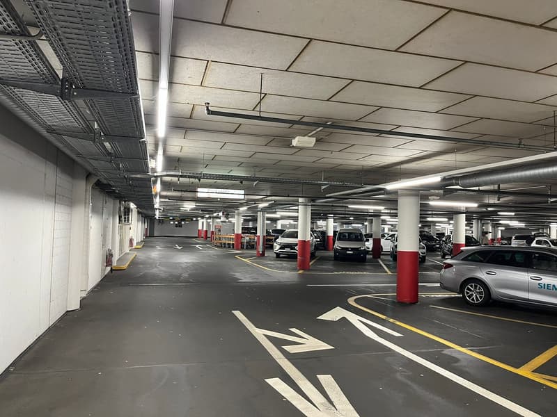 Indoor parking garage with multiple levels, concrete floors, yellow and red painted lines on the ground, and various parked cars.