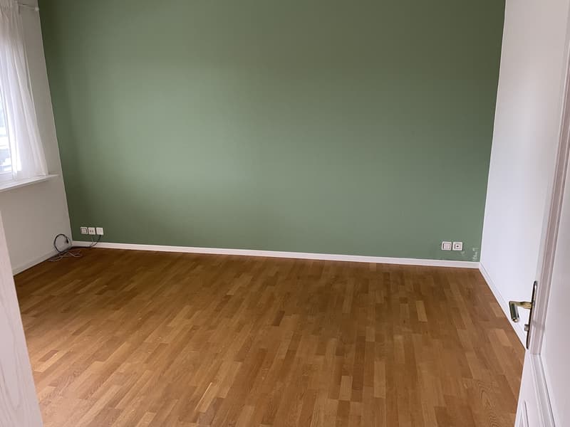 Empty room with green walls and hardwood flooring. The room appears to be a living room or bedroom with a window and electrical outlets visible.