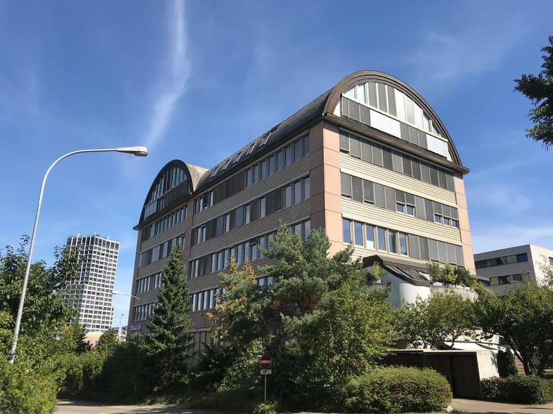 A multi-story office building with a curved, arched roof and large windows. The building has a parking area in front and is surrounded by trees and other buildings.