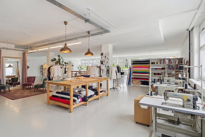 large room with white walls, wooden tables, sewing machines, shelves with books and fabrics, chairs, and a rug