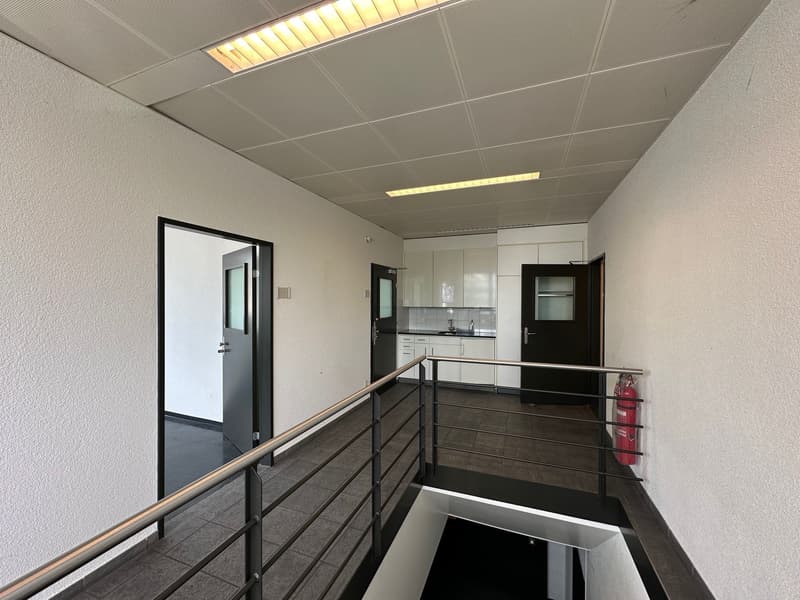 The image shows an interior office space with a kitchenette area. The space has a suspended ceiling with recessed lighting, and the walls are painted a neutral color. There is a railing along the upper level, and a door leading to another room. The kitche