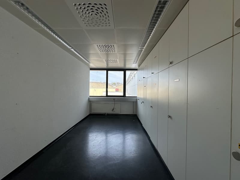 The image shows an empty office space with a suspended ceiling, windows, and lockers or storage units along one wall. The floor is a dark, smooth material, and the overall aesthetic is minimalist and functional.
