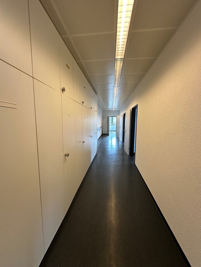 The image shows a long, narrow hallway with white walls and a dark floor. There are rows of lockers or storage units lining the walls on one side, and the hallway leads to a doorway at the far end.