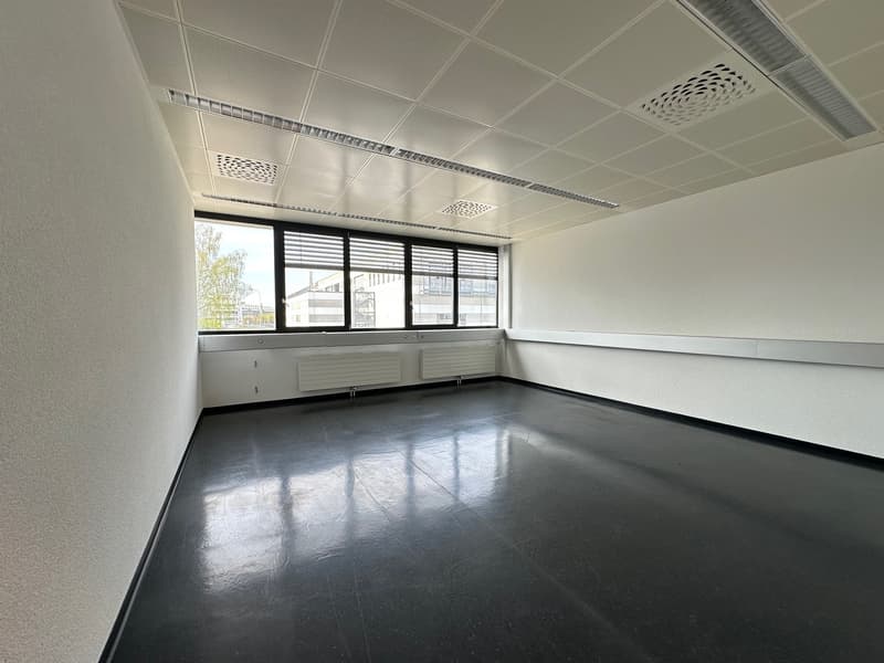 The image shows a large, open office space with a suspended ceiling, recessed lighting, and large windows providing natural light. The floor is a dark, polished concrete material. The room appears to be empty and ready for use as an office or commercial s