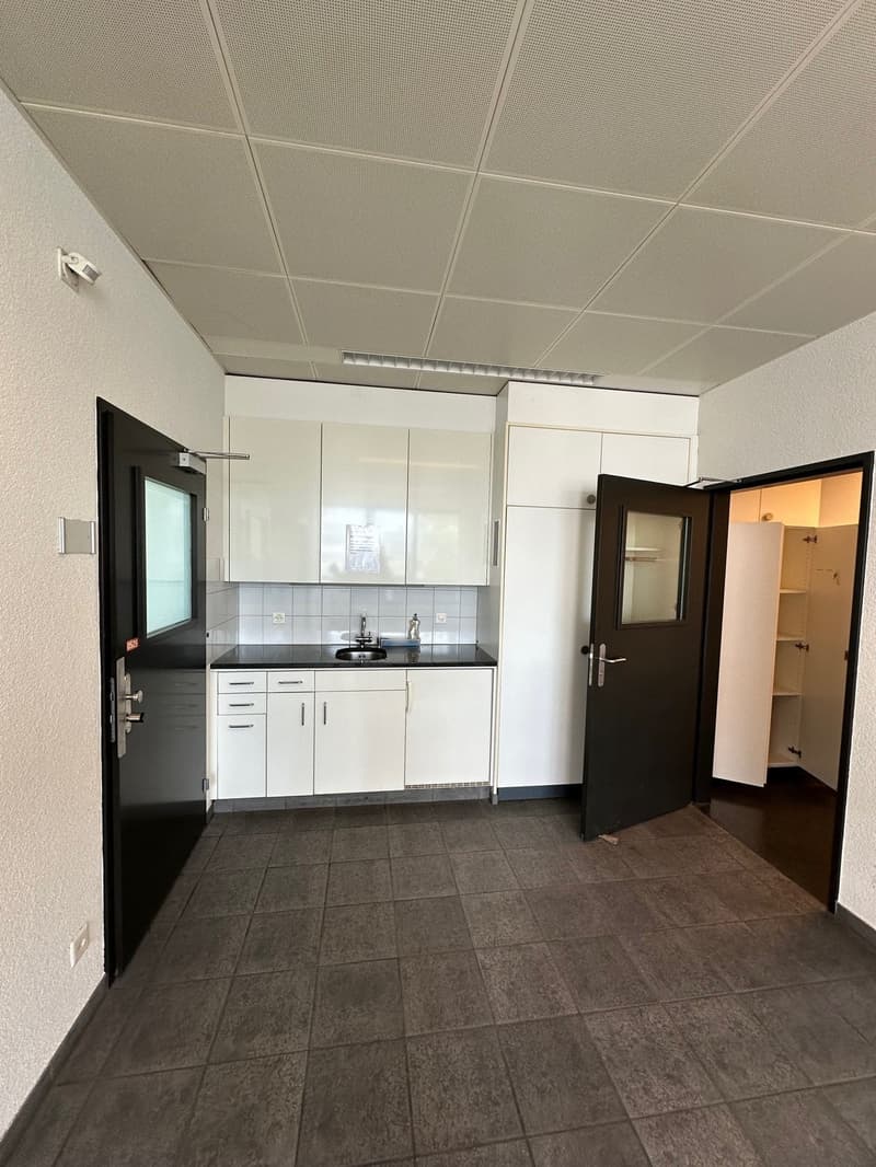 The image shows a small studio apartment with a kitchenette and a bathroom. The kitchenette has white cabinets and drawers, a black countertop, and a sink. The floor is covered with gray tile. The room has a suspended ceiling with white tiles. There are t