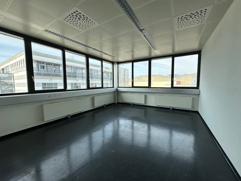 The image shows a large, open office space with floor-to-ceiling windows providing ample natural light. The room has a gray tiled floor and a suspended ceiling with circular ventilation grilles. The walls are painted a light color, and there are built-in