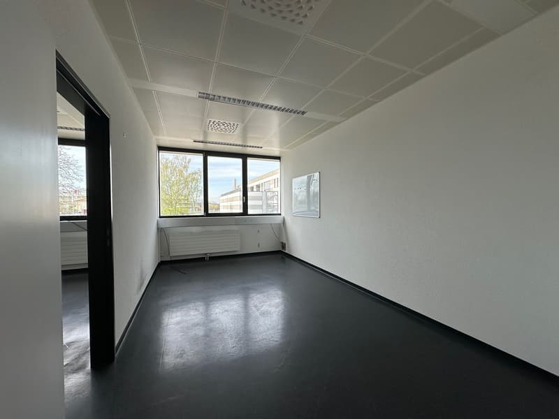 This image shows an empty office space with white walls, a suspended ceiling, and large windows overlooking the exterior. The room has a concrete floor and appears to be in a commercial or office building setting.