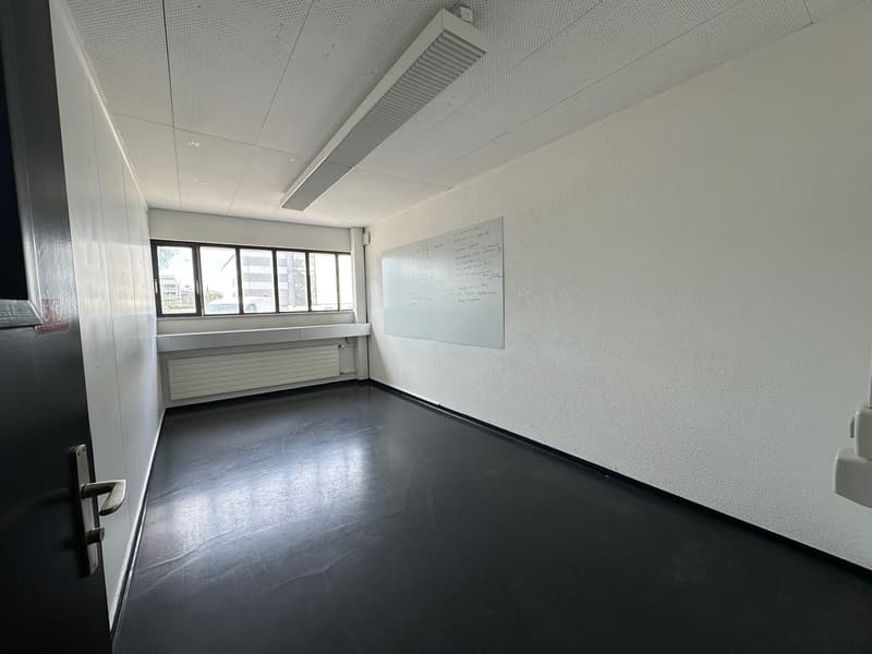 Empty office space with white walls, a whiteboard, and large windows providing natural light. The room has a concrete floor and a suspended ceiling with air conditioning vents.