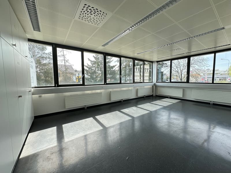 Large open office space with floor-to-ceiling windows, suspended ceiling with ventilation grilles, and concrete flooring. The space appears to be empty and ready for use.