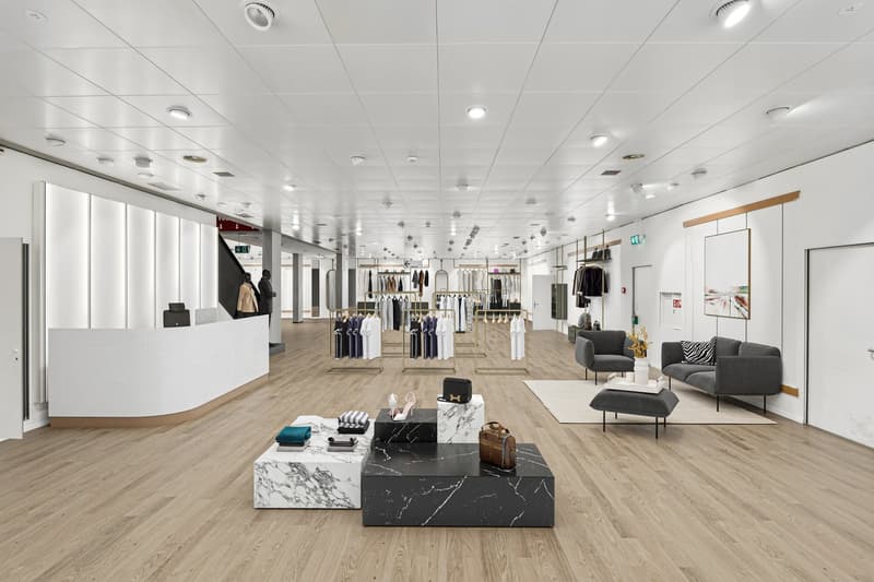 Large open retail space with high ceilings, wooden flooring, and modern furniture and decor. The space features clothing racks, display tables, and a seating area with sofas and coffee tables. The lighting is bright and uniform throughout the space.
