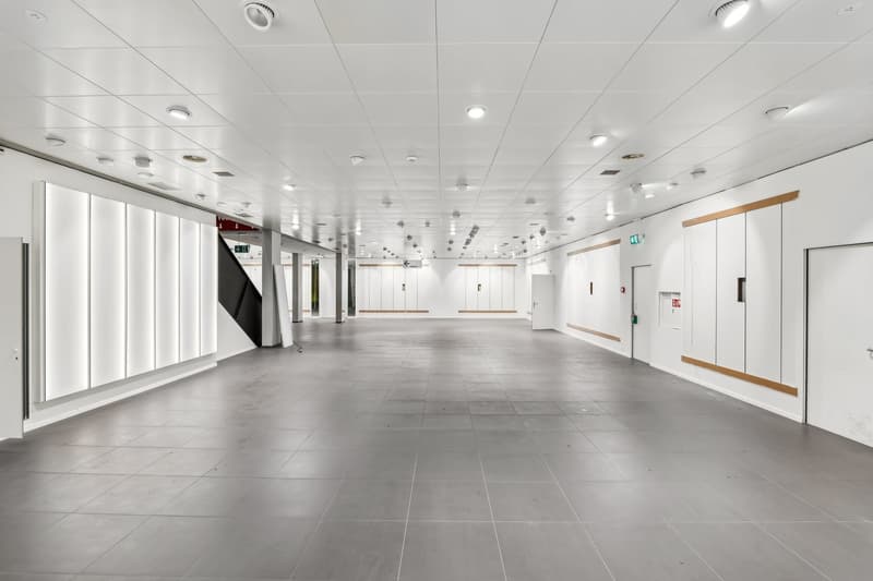 Large open commercial space with white walls, gray tile flooring, and recessed lighting throughout the ceiling. The space appears to be empty and ready for potential tenants or businesses to occupy.