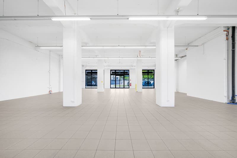 Empty room with white walls, tiled floor, and white ceiling with recessed lighting. Four white pillars support the ceiling. Three large windows with glass doors are visible in the distance.