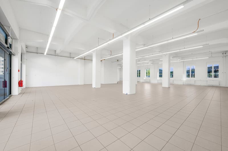 Empty room with tiled flooring, white walls, and ceiling-mounted fluorescent lights. There are white pillars and windows with white frames.