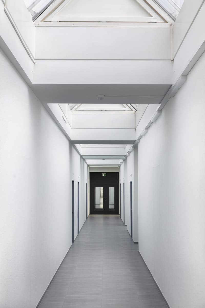 Long, narrow hallway with white walls, gray flooring, and a glass door at the end. The ceiling has a triangular skylight feature.