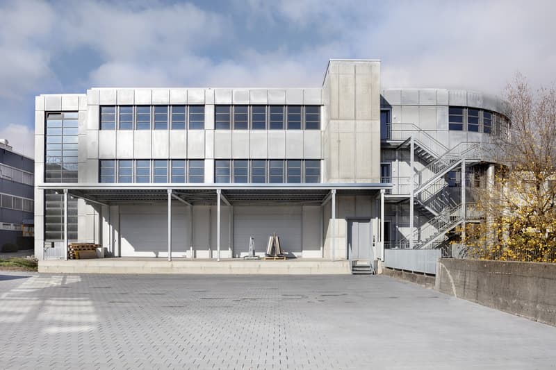 Large modern commercial building with a rectangular shape, made of concrete and glass. The building has multiple floors and a prominent entrance with stairs leading up to it. The exterior features a minimalist, industrial design with clean lines and a neu