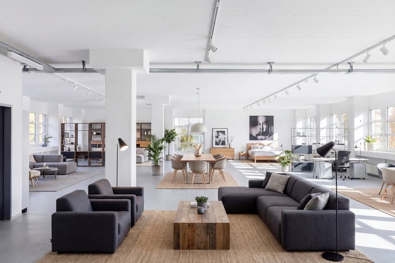 modern living room with white walls, gray couches, wooden coffee table, floor lamps, dining table, chairs, plants, bookshelves, and windows