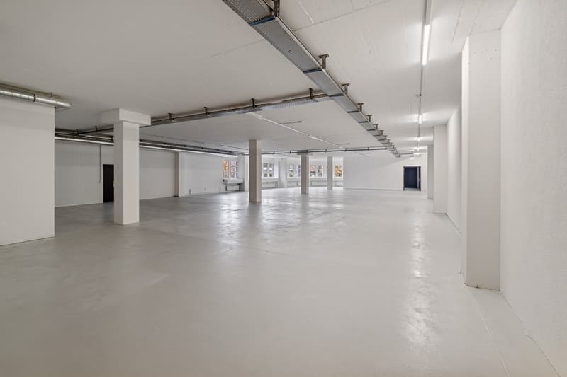 Large open floor plan with concrete flooring, white walls, and exposed ceiling with lighting fixtures. The space appears to be a commercial or industrial building, potentially suitable for office, retail, or other commercial use.