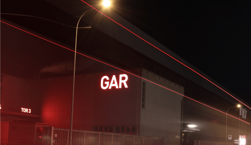 Large industrial building with the word 'GAR' illuminated in red neon lights on the facade. There are red laser-like lights visible in the night sky around the building.