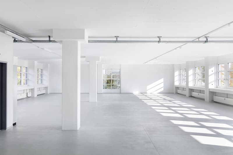 Large open-plan office space with white walls, concrete floors, and large windows allowing natural light. The space has multiple workstations and storage units along the walls.