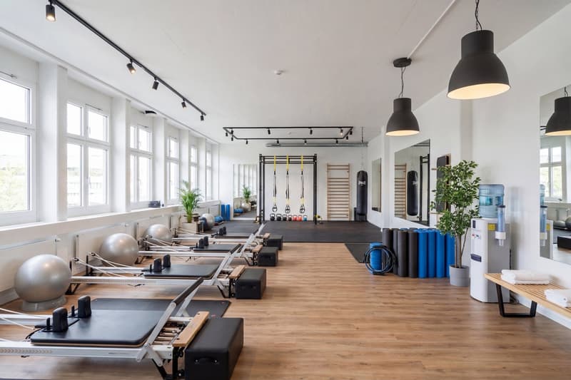 A gym with wooden floor, large windows, exercise equipment, mirrors, and plants.