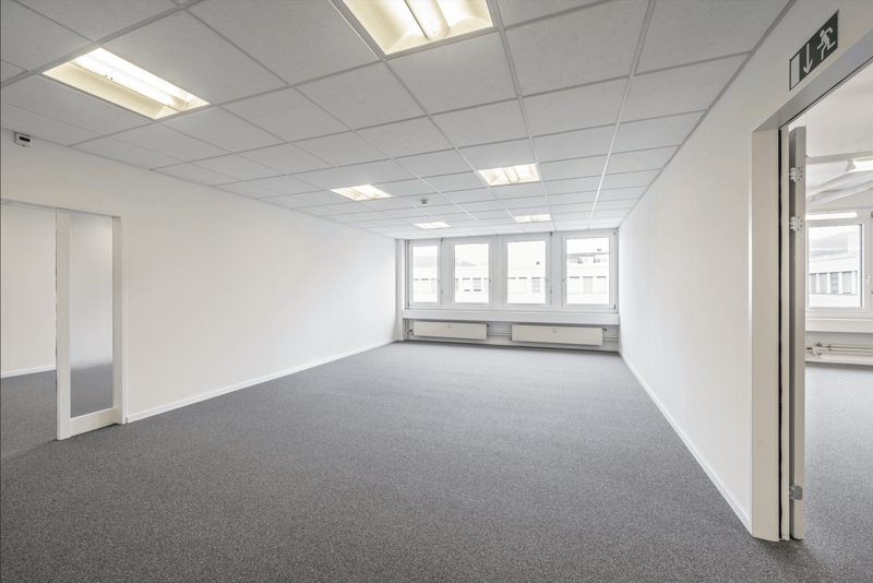 Large open office space with white walls, gray carpeted floor, and recessed lighting. The room has multiple windows providing natural light. There is a door leading to another room or area.