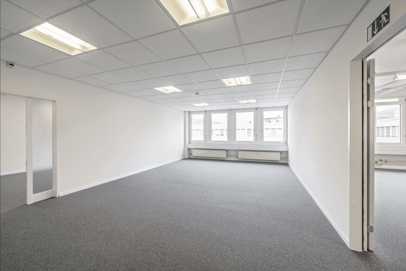 Large open office space with white walls, gray carpeted floor, and recessed lighting. The room has multiple windows providing natural light. There is a door leading to another room or area.
