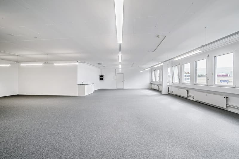 wide open room, white walls, gray floor, windows, kitchen area with a sink