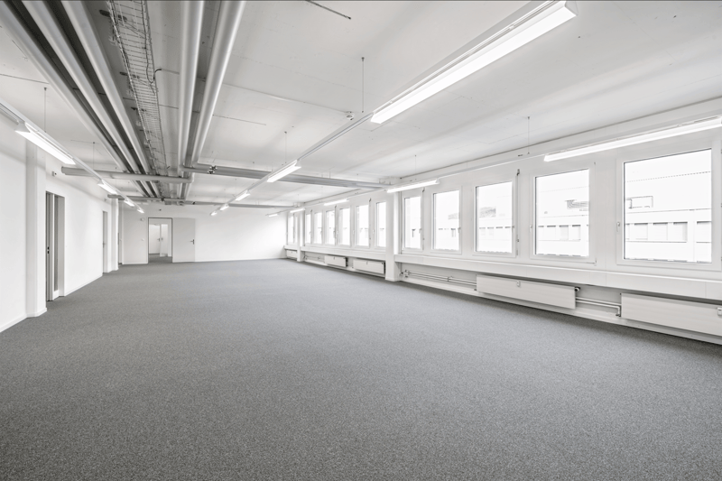 Large open office space with high ceilings, exposed ductwork, and multiple windows along the walls. The room has a gray carpeted floor and appears to be an empty commercial or office space ready for use.