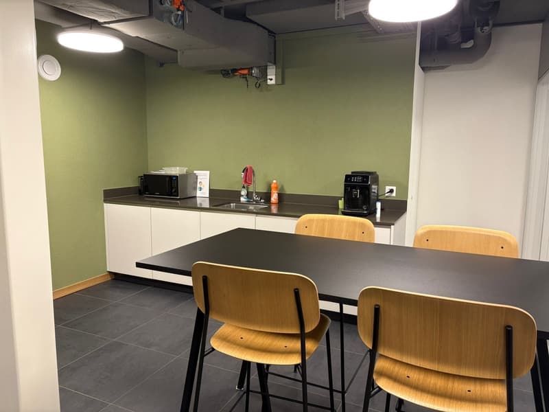 A small kitchen area with a microwave, sink, coffee machine, a large table with 4 chairs, a light on the ceiling, and green walls.