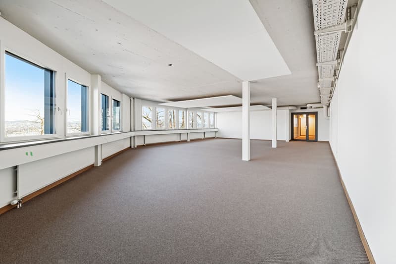 Large open-plan office space with high ceilings, white walls, and large windows providing natural light. The room has a carpeted floor and several support columns. There is a doorway visible at the far end of the room.