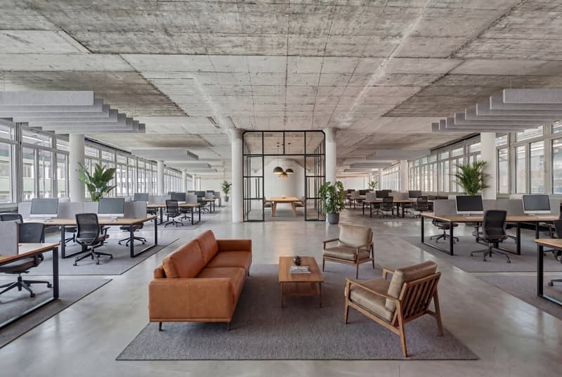 Open office layout, modern design, large windows, concrete ceilings, multiple desks and chairs, wooden coffee table and chairs, office equipment, indoor plants.