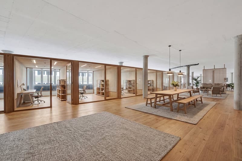 Open office space with wooden floors, glass walls, wooden desks, office chairs, wooden dining table, wooden benches, rugs, and plants.