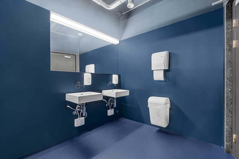Modern bathroom with blue walls, white fixtures including sinks, soap dispensers, and a hand dryer. The room also has a toilet and appears to have a dishwasher installed.