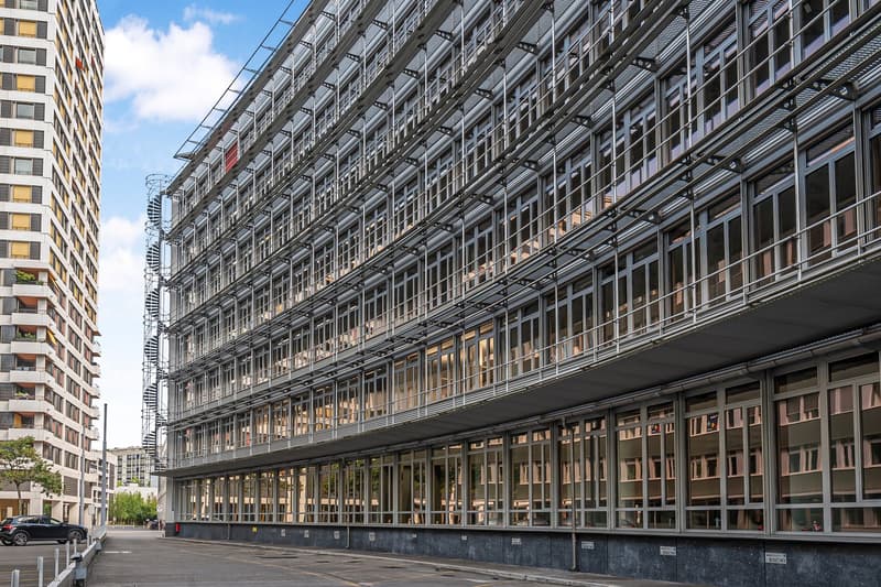 Large multi-story commercial building with a glass and steel facade, featuring numerous windows and balconies. The building appears to have a parking area or driveway in front of it.