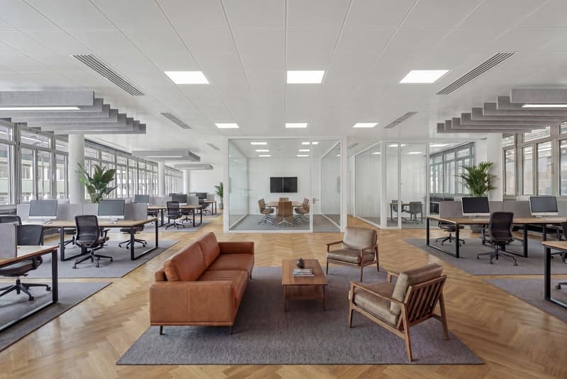 modern office layout with open-plan workspaces, large glass windows, wooden floors, office desks with computers, comfortable seating, plants, conference room with meeting table, TV monitor, and glass walls