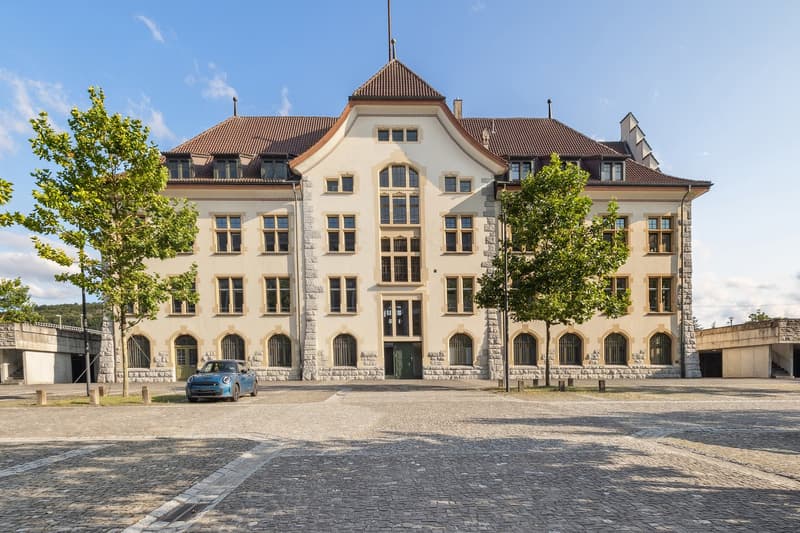 Three-story building, beige exterior, brown roof, large windows, cobblestone courtyard, tree in front