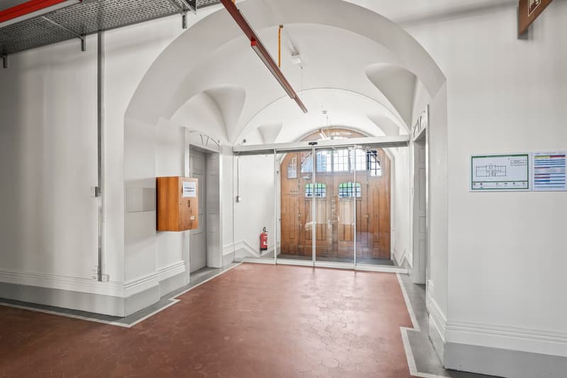 Interior hallway, red tiled floor, arched ceiling, two doors, one leading to another hallway with wooden doors and windows, fire extinguisher, information poster.